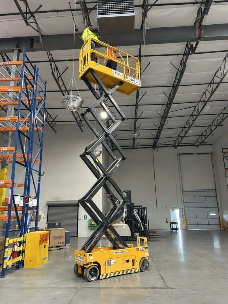 HVAC technicians install a commercial air conditioning system inside a warehouse facility in Kingman, Arizona using a yellow scissor lift near ceiling-mounted ventilation equipment. The image shows commercial ductwork, industrial shelving, warehouse lighting, polished concrete flooring, and technicians wearing safety equipment during a commercial AC installation project. Ambient Edge provides commercial air conditioning installation services in Kingman, AZ for warehouses, distribution centers, industrial buildings, and commercial facilities that require reliable indoor cooling systems. Business owners in Kingman can contact Ambient Edge at 928-615-8224 for commercial HVAC installation, warehouse cooling systems, rooftop AC services, and large-scale commercial air conditioning projects.